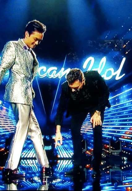 Two presenters on a TV stage, one in a silver jacket and dark leather shoes